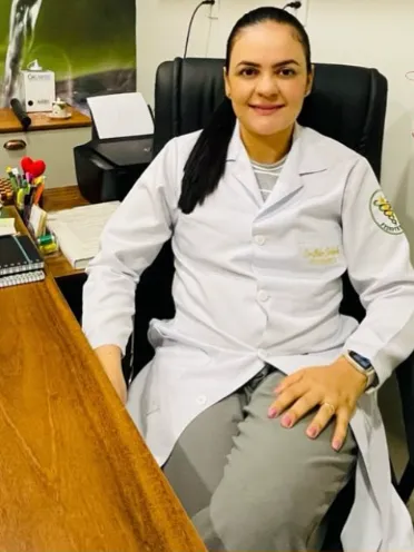 Luiza, de jaleco branco, sentada à mesa do consultório, sorrindo